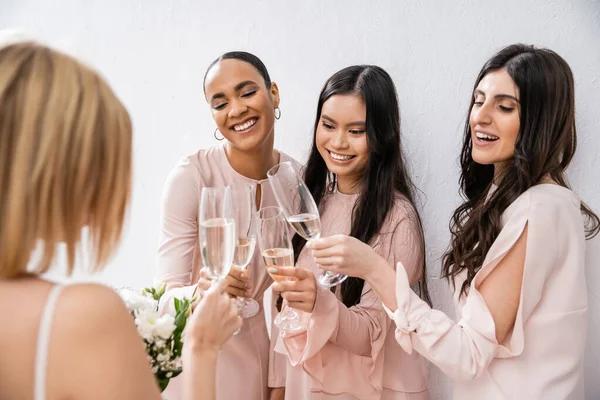 Namoradas multiculturais alegres copos clinking com champanhe, noiva com flores brancas, mulheres morenas e loiras, damas de honra, diversidade, positividade, buquê de noiva, fundo cinza — Fotografia de Stock