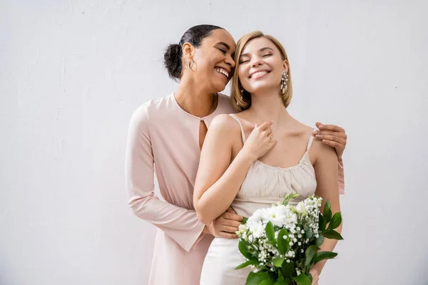 Special occasion, cheerful bride with bridesmaid, happy interracial women, wedding dress and bridesmaid gown, african american woman hugging engaged friend on grey background — Stock Photo