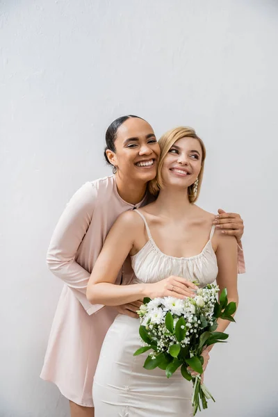 Bridal party, happy bride with bridesmaid, cheerful interracial women, wedding dress and bridesmaid gown, african american woman hugging engaged friend on grey background — Stock Photo