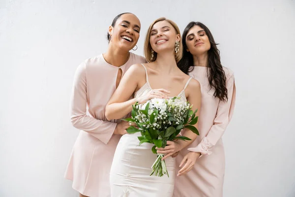 Bridal party, special occasion, brunette and interracial bridesmaids hugging blonde bride, friendship goals, grey background, happy multicultural girlfriends, cultural diversity, bridal bouquet — Stock Photo