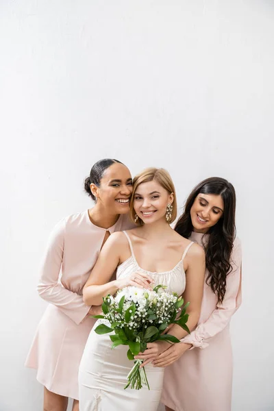 Bridal party, special occasion, brunette and interracial bridesmaids hugging blonde bride, friendship goals, grey background, cheerful multicultural girlfriends, cultural diversity, bridal bouquet — Stock Photo