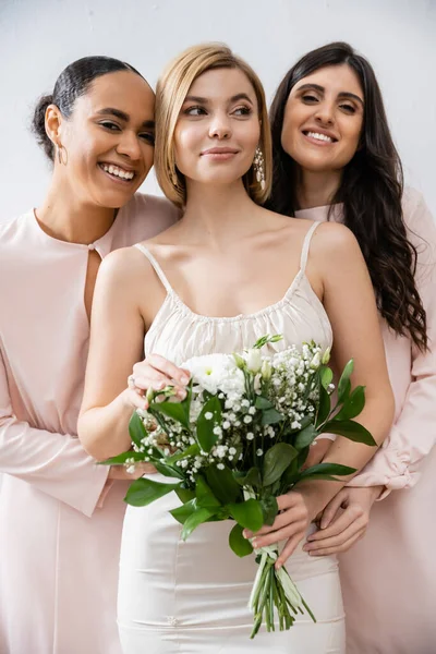Special occasion, brunette and interracial bridesmaids hugging blonde bride, friendship goals, grey background, happy multicultural girlfriends, cultural diversity, bridal bouquet, bridal party — Stock Photo