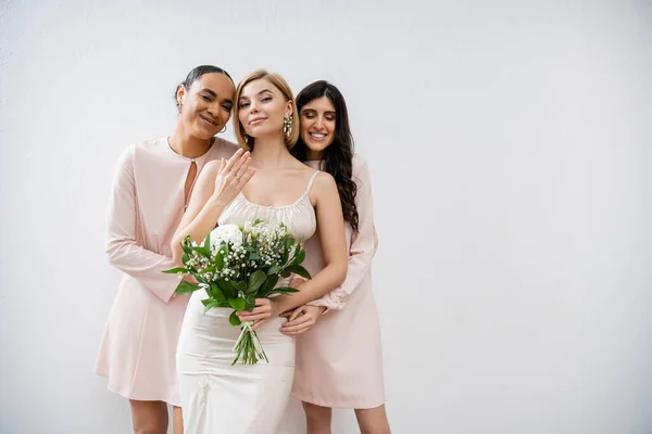 Special occasion, bridesmaids hugging bride, friendship goals, grey background, happy multicultural girlfriends, cultural diversity, bridal bouquet, bridal party, blonde and brunette women — Stock Photo