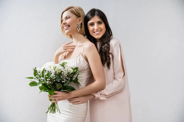 Special occasion, bridesmaid hugging bride, friendship goals, grey background, cheerful girlfriends, bridal bouquet, blonde and brunette women, white flowers, happiness — Stock Photo