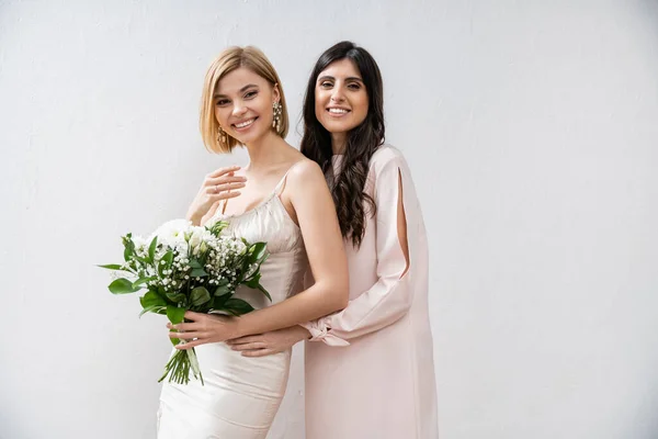 Special occasion, cheerful bridesmaid hugging bride, friendship goals, grey background, happy girlfriends, bridal bouquet, blonde and brunette women, white flowers, happiness — Stock Photo