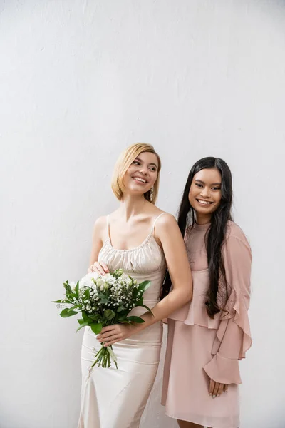 Special occasion, asian bridesmaid smiling near bride, friendship goals, grey background, happy girlfriends, bridal bouquet, blonde and brunette women, white flowers, happiness — Stock Photo