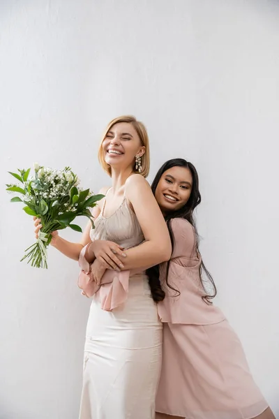 Special occasion, asian bridesmaid hugging beautiful bride, friendship goals, grey background, happy girlfriends, bridal bouquet, blonde and brunette women, white flowers, happiness — Stock Photo