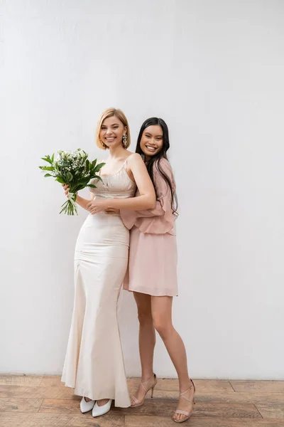 Special occasion, cheerful asian bridesmaid hugging beautiful bride, friendship goals, grey background, happy girlfriends, bridal bouquet, blonde and brunette women, white flowers, happiness — Stock Photo