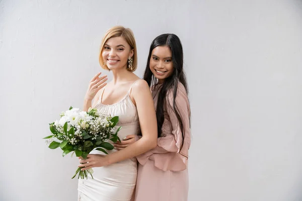 Special occasion, asian bridesmaid hugging beautiful bride, friendship goals, grey background, happy girlfriends, bridal bouquet, blonde and brunette women, white flowers, positivity — Stock Photo