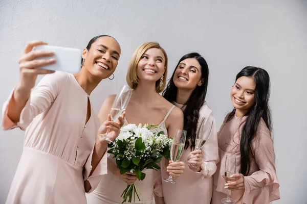 Quatre femmes, mariée gaie et ses demoiselles d'honneur multiculturelles prenant selfie ensemble, bonheur, verres de champagne, bouquet de mariée, robe de mariée, femmes brunes et blondes — Photo de stock