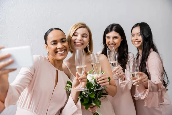 Четыре женщины, делающие селфи, веселая невеста и ее межрасовые подружки невесты, счастье, бокалы для шампанского, букет невесты, свадебное платье, платье подружки невесты, брюнетка и блондинка — стоковое фото