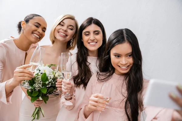 Quattro donne, sposa bionda positiva e le sue damigelle d'onore interrazziale prendendo selfie insieme, bicchieri di champagne, bouquet da sposa, abito da sposa, abito da damigella d'onore, donne brune e bionde — Foto stock