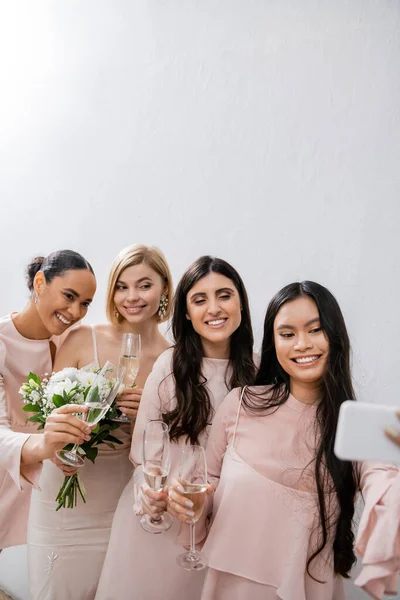Quatro mulheres, noiva loira alegre e suas damas de honra inter-raciais tomando selfie juntos, felicidade, copos de champanhe, buquê de noiva, vestido de noiva, vestido de dama de honra, morena e mulheres loiras — Fotografia de Stock