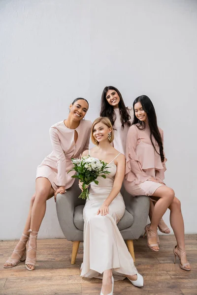 Свадебная фотография, четыре женщины, подружки невесты и невесты, межрасовые подруги, день свадьбы, культурное разнообразие, сидя на кресле, серый фон, счастье и радость, свадебное платье — стоковое фото