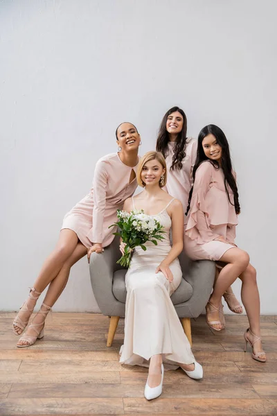 Свадебная фотография, счастливые четыре женщины, подружки невесты и невесты, межрасовые подруги, день свадьбы, культурное разнообразие, сидя на кресле, серый фон, счастье и радость, свадебное платье — стоковое фото