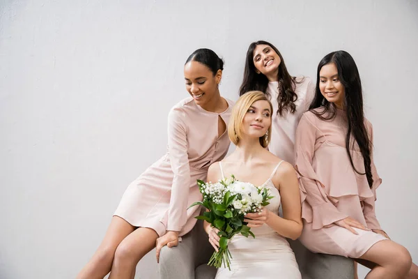 Свадебная фотография, четыре женщины, радостные подружки невесты и невесты, межрасовые подружки, день свадьбы, культурное разнообразие, сидение на кресле, серый фон, счастье и радость, свадебное платье — стоковое фото