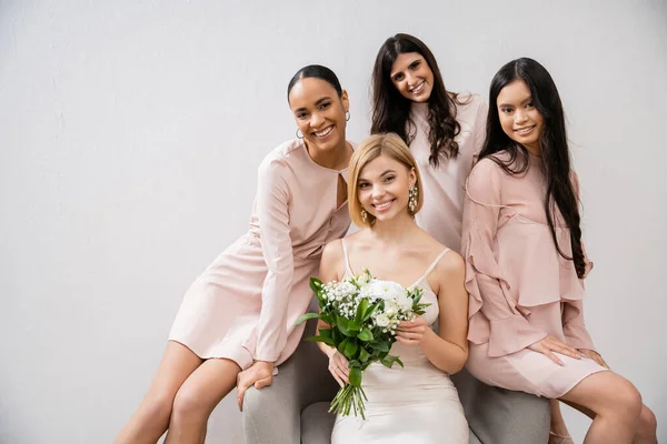 Wedding photography, cultural diversity, four women, joyful bride with bouquet and bridesmaids, interracial, wedding day, sitting on armchair, grey background, happiness and joy, wedding dress — Stock Photo
