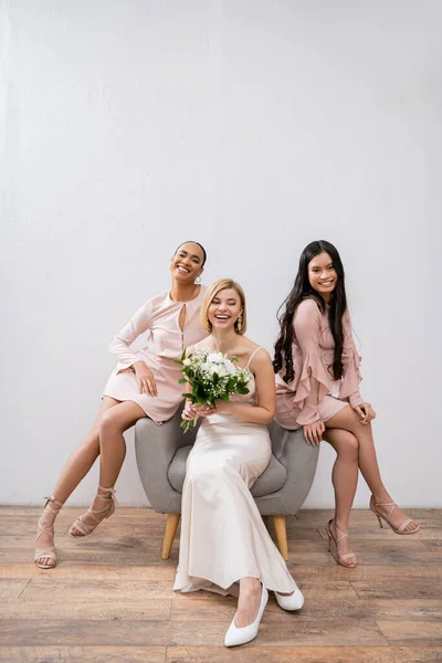 Wedding photography, cultural diversity, three women, happy bride with bouquet and her interracial bridesmaids sitting on armchair on grey background, brunette and blonde, joy, celebration — Stock Photo
