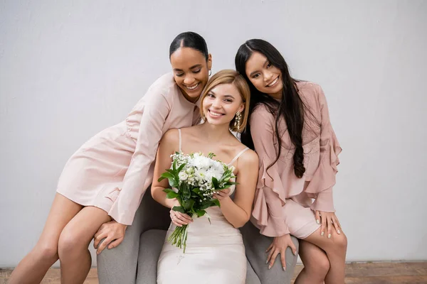 Свадебная фотография, разнообразие, три женщины, счастливая невеста с букетом и ее межрасовые подружки невесты, сидящие на кресле на сером фоне, брюнетка и блондинка, радость, праздник — стоковое фото