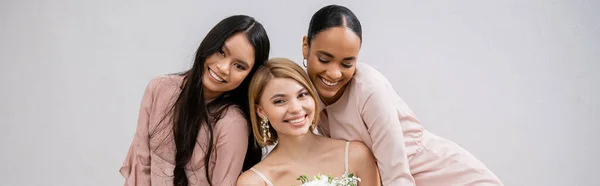 Свадебная фотография, культурное разнообразие, три женщины, счастливая невеста с букетом и ее межрасовые подружки невесты, сидящие на кресле на сером фоне, брюнетка и блондинка, радость, праздник, баннер — стоковое фото