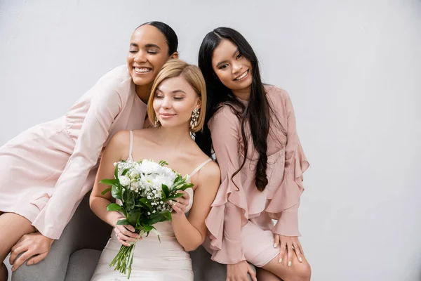Wedding theme, cultural diversity, three women, cheerful bride with bouquet and her interracial bridesmaids sitting on armchair on grey background, brunette and blonde, joy, celebration — Stock Photo