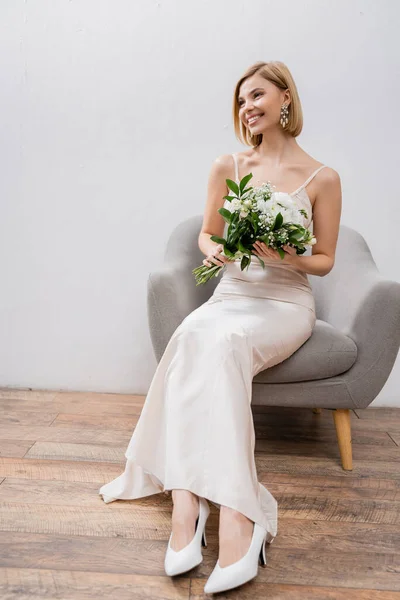 Wedding photography, special occasion, beautiful, blonde bride in wedding dress sitting in armchair and holding bouquet on grey background, white flowers, bridal accessories, happiness — Stock Photo