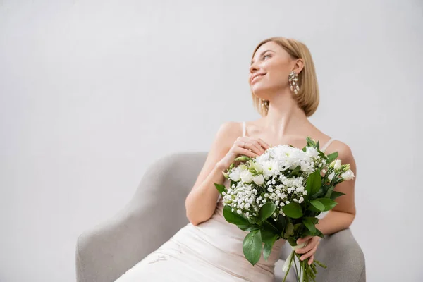 Special occasion, happy blonde bride in wedding dress sitting in armchair and holding bouquet on grey background, engagement ring, white flowers, bridal accessories, happiness, feminine — Stock Photo