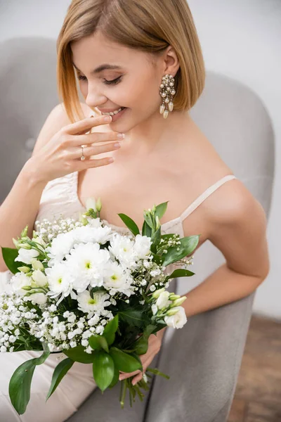 Besonderer Anlass, wunderschöne blonde Braut im Hochzeitskleid sitzt im Sessel und hält Strauß auf grauem Hintergrund, Verlobungsring, weiße Blumen, Brautzubehör, Glück, feminin — Stockfoto