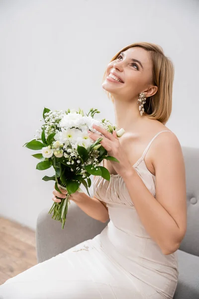 Attractive and blonde bride in wedding dress sitting in armchair and holding bouquet on grey background, white flowers, bridal accessories, happiness, special occasion, smiling, feminine, blissful — Stock Photo