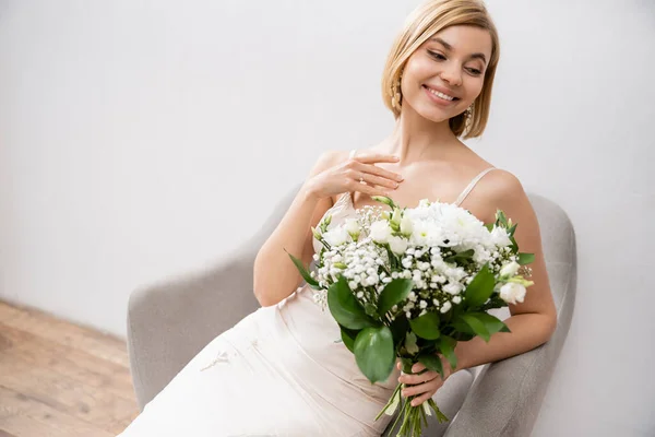 Fröhliche und elegante Braut im Hochzeitskleid sitzt im Sessel und hält Blumenstrauß auf grauem Hintergrund, weiße Blumen, Brautzubehör, Glück, besonderen Anlass, schön, feminin, selig — Stockfoto