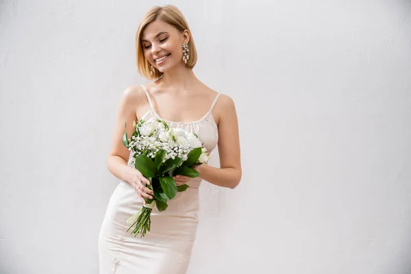 Freudige und blonde Braut im Brautkleid mit Blumenstrauß auf grauem Hintergrund, weiße Blumen, Brautzubehör, Glück, besonderer Anlass, schön, feminin, selig — Stockfoto