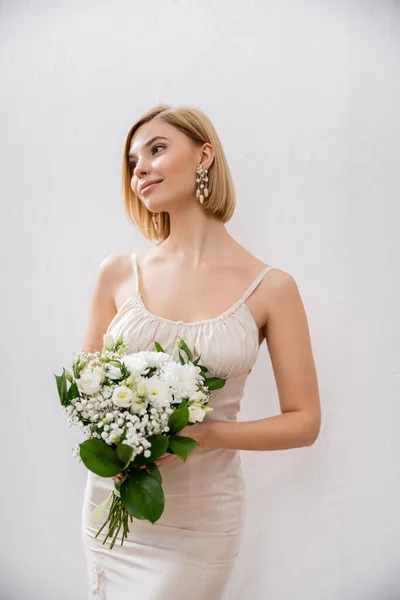 Attractive and blonde bride in wedding dress holding bouquet on grey background, white flowers, bridal accessories, happiness, special occasion,   beautiful, feminine, blissful — Stock Photo