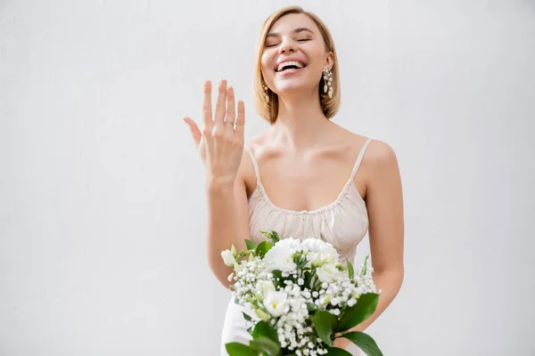 Особливий випадок, чудова блондинка наречена у весільній сукні тримає букет і показує заручини кільце, білі квіти, весільні аксесуари, щастя, сірий фон — стокове фото