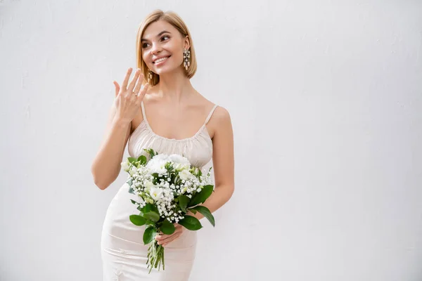 Свадебная тема, великолепная блондинка невеста в свадебном платье проведение букет и показывая обручальное кольцо, белые цветы, свадебные аксессуары, счастье, серый фон, особый случай — стоковое фото