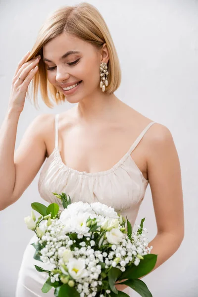 Cheerful and blonde bride in wedding dress holding bouquet on grey background, white flowers, bridal accessories, happiness, special occasion,   beautiful, feminine, blissful — Stock Photo