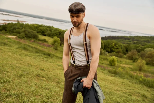 Стильный бородатый мужчина в кепке газетчика и подтяжках, держась за руку в кармане брюк и пиджака, глядя в камеру и стоя с пейзажем на заднем плане, мода вперед в сельской местности — стоковое фото