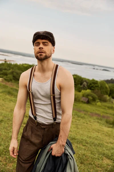Portrait of fashionable and bearded man in suspenders and newsboy cap holding jacket and looking at camera while standing with blurred landscape at background, fashion-forward in countryside — Stock Photo