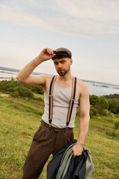 Trendy bearded man in suspenders touching newsboy cap and holding jacket while looking at camera with rural landscape at background, fashion-forward in countryside — Stock Photo
