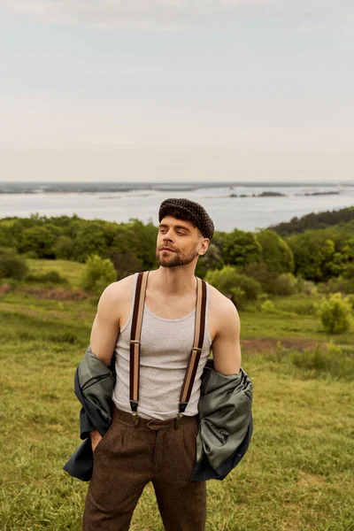 Fashionable and bearded man in jacket, suspenders and newsboy cap holding hands in pockets and looking away while standing with blurred nature at background, fashion-forward in countryside — Stock Photo