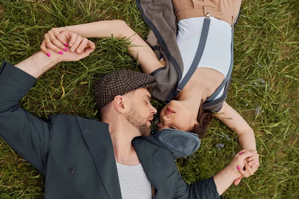Top view of trendy and bearded man in newsboy cap and vintage jacket holding hands of stylish girlfriend in susenders while lying on grassy lawn, fashion-forward in countryside — стоковое фото