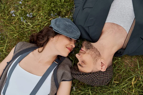 Top view of smiling and fashionable woman in newsboy cap lying with closed eyes near bearded boyfriend in jacket and vintage outfit on grassy lawn, fashion-forward in countryside — Stock Photo
