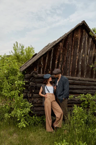 Fashionable woman in vintage outfit touching suspender while standing and looking at bearded boyfriend in newsboy cap near rural house on meadow, fashion-forward in countryside — Stock Photo