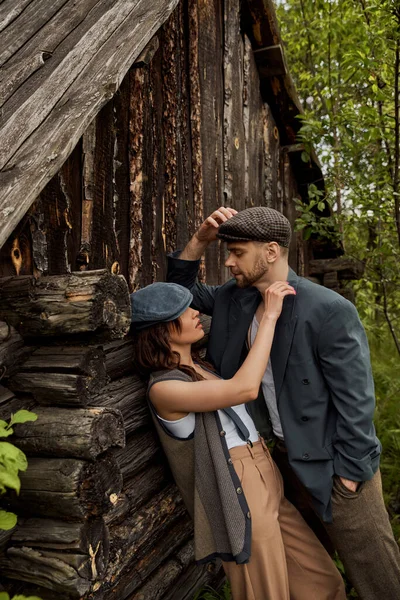 Fashionable woman in vintage vest and suspenders touching shoulder of bearded boyfriend in jacket and newsboy cap while standing near rural house, stylish couple in rural setting — Stock Photo