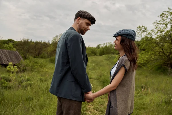 Bearded and stylish man in jacket and newsboy cap holding hand and looking at cheerful girlfriend in vest and standing together on grassy lawn at overcast, stylish couple in rural setting — Stock Photo