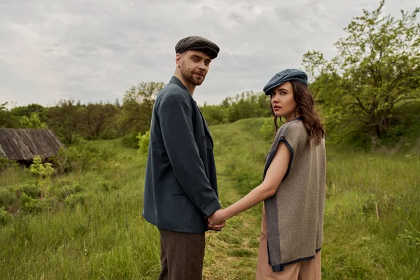 Fashionable woman in vest and newsboy cap looking at camera and holding hand of bearded boyfriend in jacket and standing with landscape and overcast at background, stylish couple in rural setting — Stock Photo