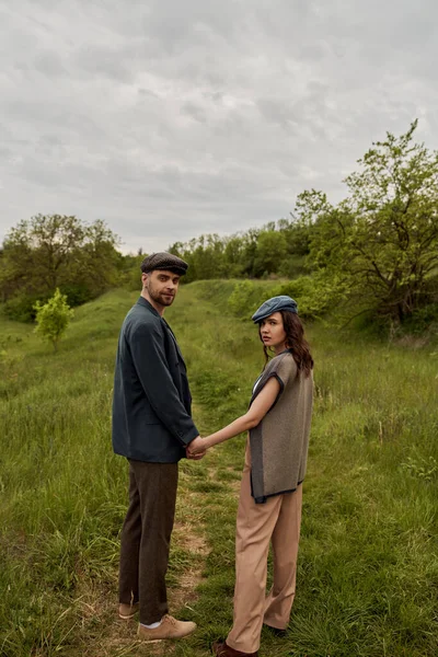 Fashionable bearded man in jacket and newsboy cap holding hand of brunette girlfriend and looking at camera with landscape and overcast at background, stylish couple in rural setting — Stock Photo