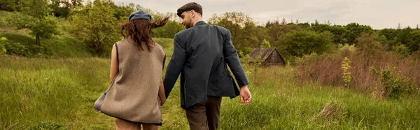 Hombre de moda y barbudo con chaqueta y gorra de reportero tomados de la mano de la novia morena en chaleco mientras caminan juntos en el prado con la naturaleza en el fondo, pareja elegante en el entorno rural, pancarta — Stock Photo