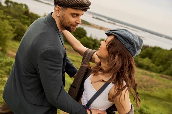 Cheerful and fashionable brunette woman in vintage suspenders and vest hugging bearded boyfriend in jacket and newsboy cap with landscape at background, fashion-forwards in countryside — Stock Photo