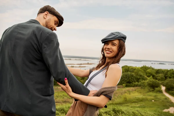 Cheerful brunette woman in vest and newsboy cap touching bearded boyfriend in jacket and looking away with nature and overcast sky at background, fashion-forwards in countryside — Stock Photo