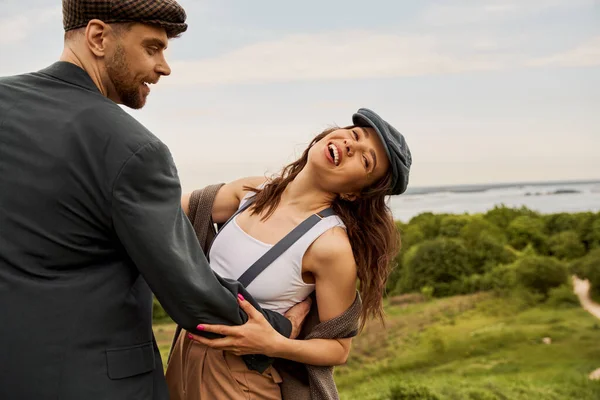 Smiling and bearded man in jacket and newsboy cap hugging cheerful girlfriend in vest and suspenders while standing with nature and sky at background, fashion-forwards in countryside — Stock Photo